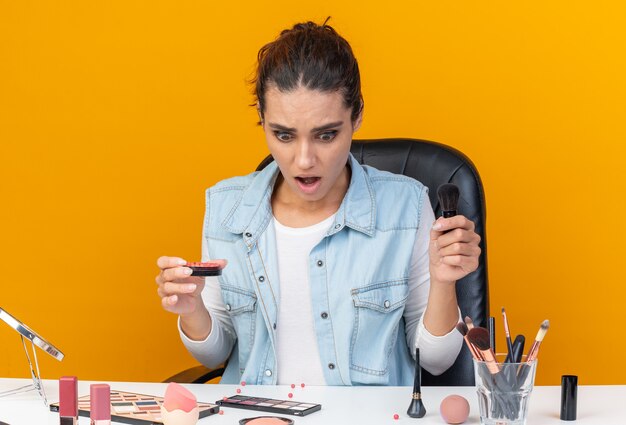 Anxious pretty caucasian woman sitting at table with makeup tools holding makeup brush and blush looking at table 