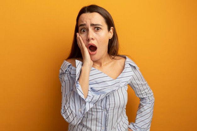 Anxious pretty woman puts hand on face isolated on orange wall