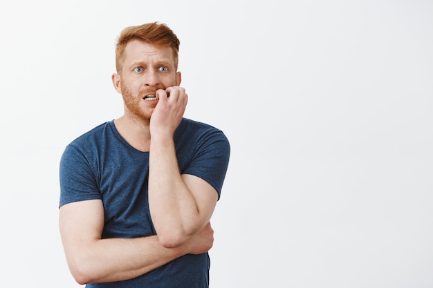 Anxious or worried handsome mature guy with red hair and bristle, biting fingernails nervous and gazing aside, standing over gray wall impatient