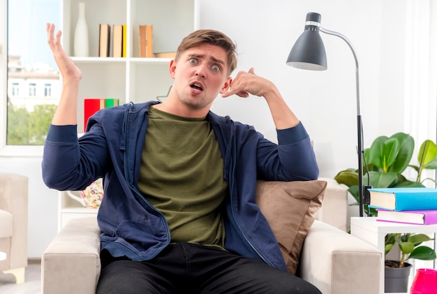 Free photo anxious young blonde handsome man sits on armchair gesturing phone hand sign and raising hand inside the living room