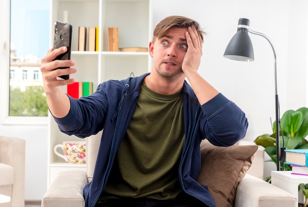 Free photo anxious young blonde handsome man sits on armchair looking at phone putting hand