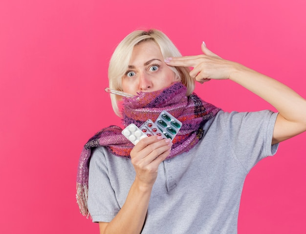 Free photo anxious young blonde ill slavic woman wearing scarf gestures gun hand sign putting at temple holds packs of medical pills measuring temperature with thermometer  on pink wall