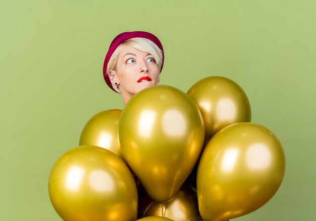 Anxious young blonde party girl wearing party hat standing behind balloons looking at side biting lip isolated on olive green background