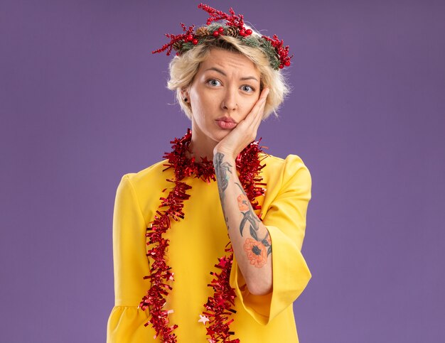 Free Photo anxious young blonde woman wearing christmas head wreath and tinsel garland around neck keeping hand on face looking  isolated on purple wall with copy space