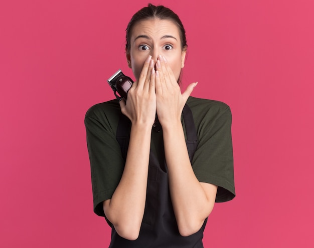 Anxious young brunette barber girl in uniform puts hands on mouth holding hair clippers on pink