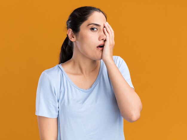 Anxious young brunette caucasian girl puts hand on face and looks at camera on orange