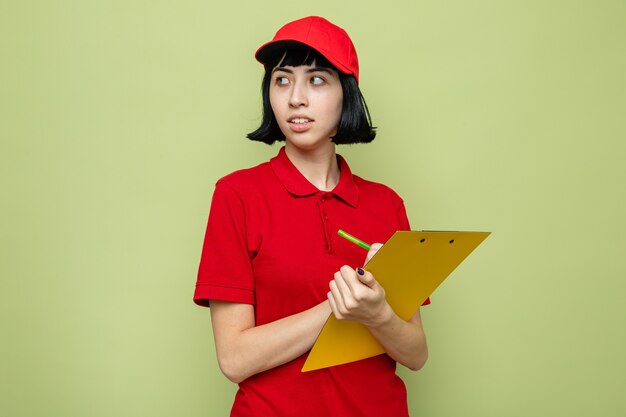 Anxious young caucasian delivery girl holding clipboard and pen looking at side 