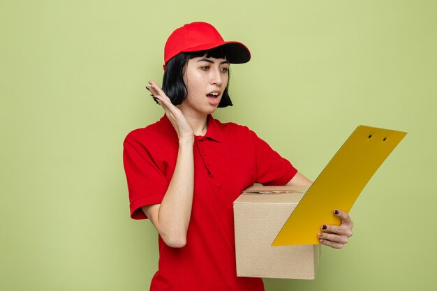 Anxious young caucasian delivery woman holding cardboard box and looking at clipboard 