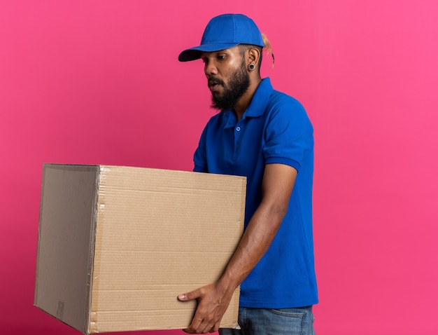 anxious young delivery man holding heavy cardboard box isolated on pink wall with copy space