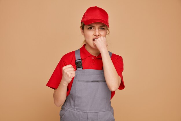 Anxious young female construction worker wearing cap and uniform clenching fist biting another fist 