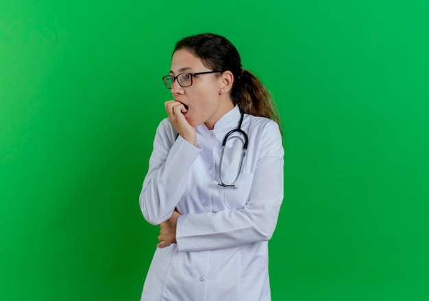 Anxious young female doctor wearing medical robe and stethoscope and glasses looking at side biting fingers isolated on green wall with copy space