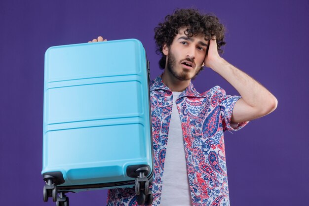 Free Photo anxious young handsome curly traveler man holding suitcase with hand on head on isolated purple space