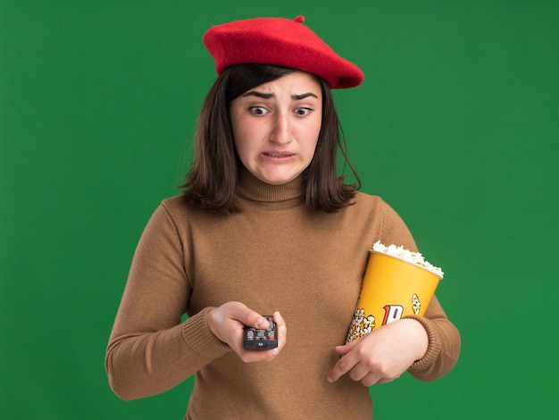 Free Photo anxious young pretty caucasian girl with beret hat holding tv controller and popcorn bucket