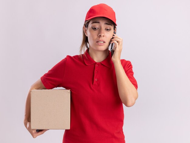 Anxious young pretty delivery woman in uniform holds cardbox and talks on phone isolated on white wall