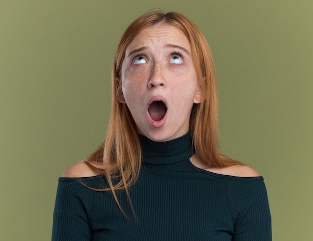 Anxious young redhead ginger girl with freckles looking up isolated on olive green wall with copy space