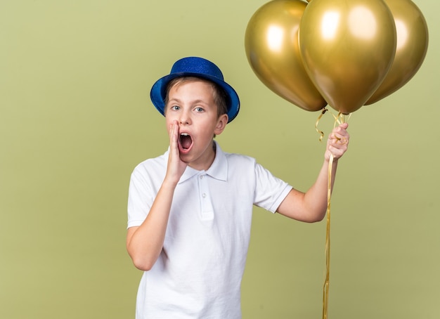 Free Photo anxious young slavic boy with blue party hat holding helium balloons and keeping hand close to mouth calling someone isolated on olive green wall with copy space