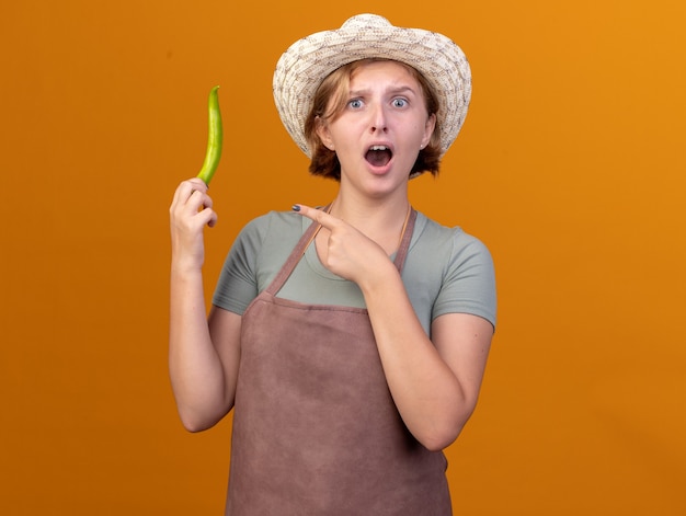 Anxious young slavic female gardener wearing gardening hat holding and pointing at hot pepper