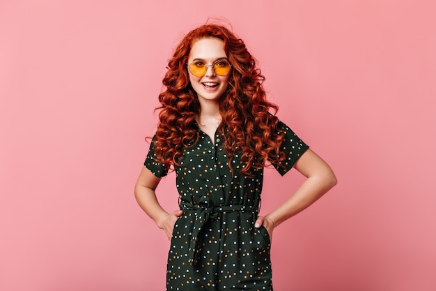 Free photo appealing ginger girl standing with hands on hips. happy curly young woman in sunglasses posing on pink background.