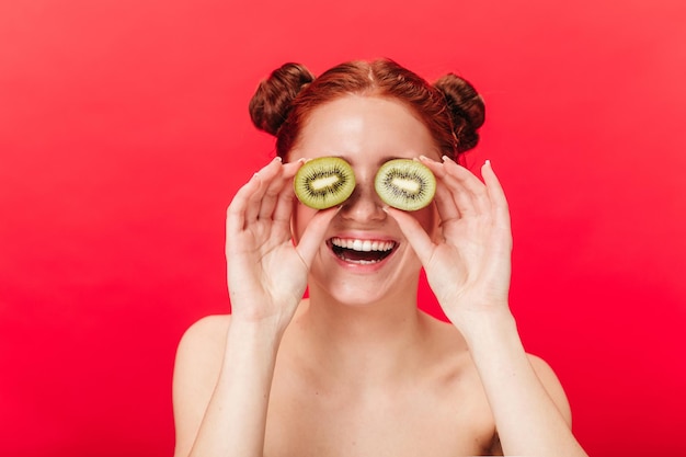 Free photo appealing girl with kiwi laughing at camera studio shot of nude ginger woman with tropical fruits isolated on red background