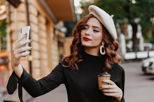 Free photo appealing long-haired girl using phone for selfie on the street. amazing ginger woman enjoying coffee outdoor.