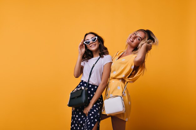 Appealing young ladies dancing together. Indoor portrait of cheerful sisters in trendy attire standing on yellow.