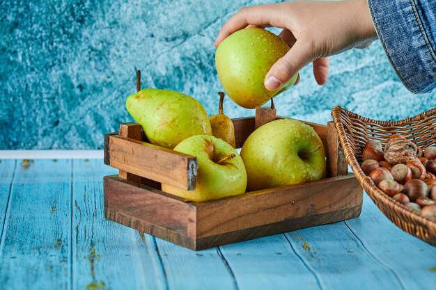 Apples and pear in wooden basket and bowl of hazelnuts on blue surface.