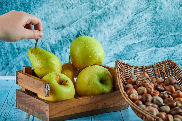 Apples and pear in wooden basket and bowl of hazelnuts on blue table.