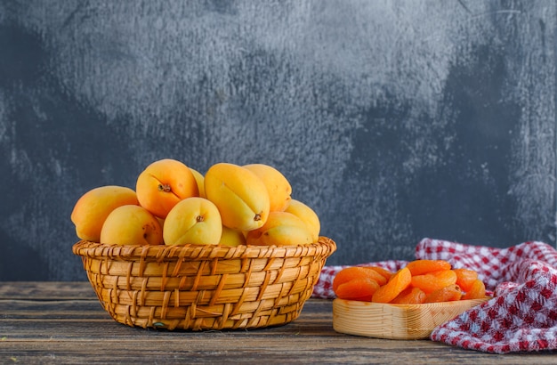 Free photo apricots with dried apricots, picnic cloth in a wicker basket on plaster and wooden table, side view.