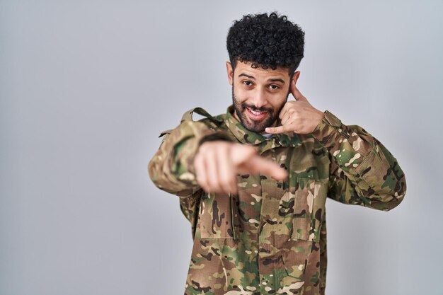 Arab man wearing camouflage army uniform smiling doing talking on the telephone gesture and pointing to you. call me.