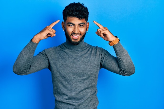 Arab man with beard wearing turtleneck sweater smiling pointing to head with both hands finger, great idea or thought, good memory