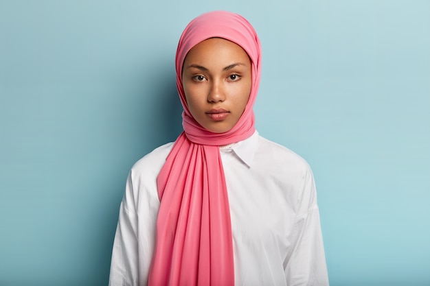 Free photo arabian woman with calm serious expression, has dark skin, wrapped in pink veil, wears white shirt, has no make up, natural beauty, models over blue wall. close up shot of muslim lady