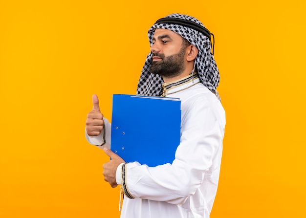 Free photo arabic businessman in traditional wear holding blue folder showing thumbs up standing sideways over orange wall