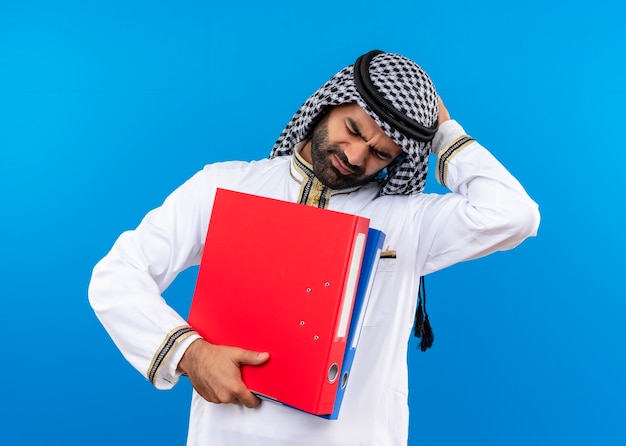 Free photo arabic businessman in traditional wear holding two folders looking confused and very anxious standing over blue wall