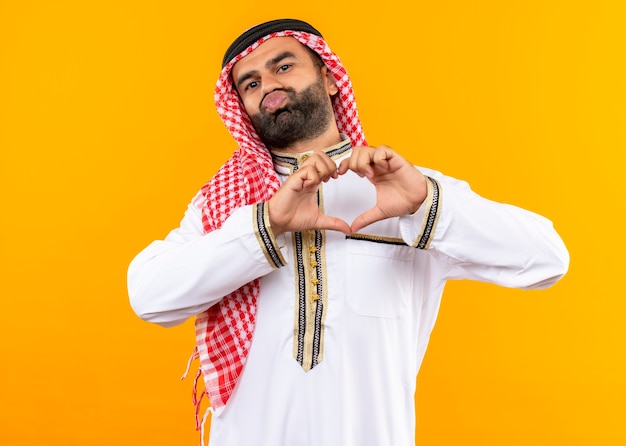 Free Photo arabic businessman in traditional wear making heart gesture with fingers over chest trying to give a kiss standing over orange wall