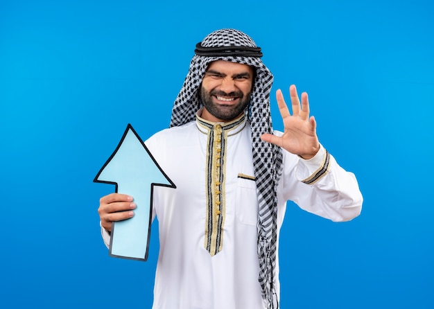 Free photo arabic man in traditional wear holding big blue arrow with annoyed expression on face standing over blue wall