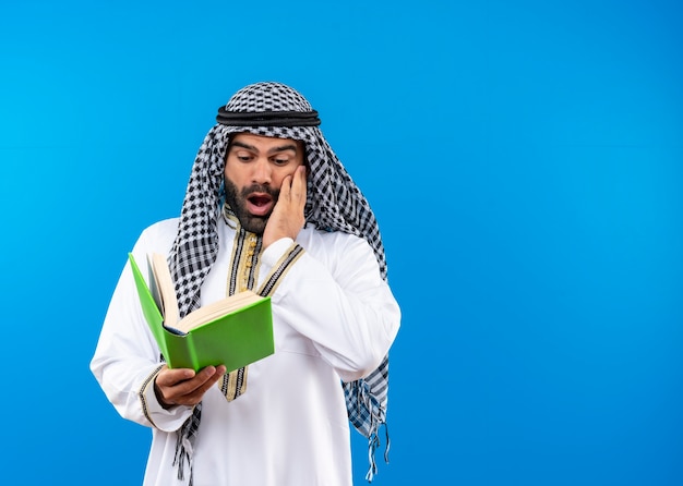 Free photo arabic man in traditional wear looking in a book shocked standing over blue wall