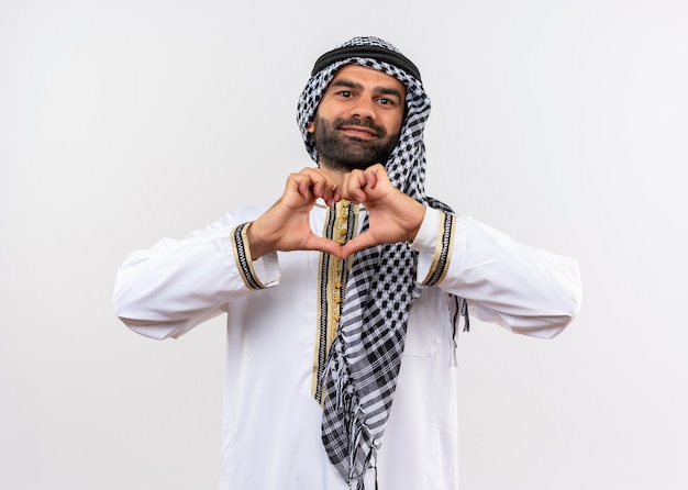 Arabic man in traditional wear making heart gesture with fingers smiling happy and positive standing over white wall