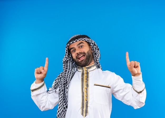 Free Photo arabic man in traditional wear pointing with fingers up smiling cheerfully standing over blue wall