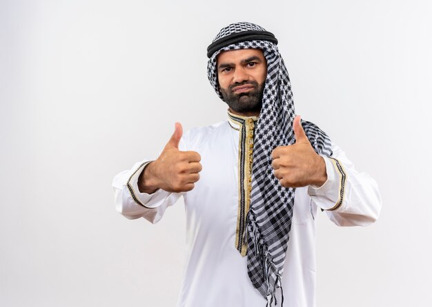 Arabic man in traditional wear smiling showing thumbs up standing over white wall