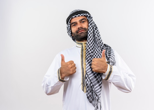 Arabic man in traditional wear smiling showing thumbs up standing over white wall