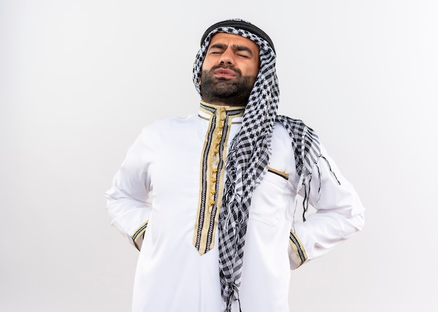 Free photo arabic man in traditional wear touching back looking unwell having pain standing over white wall