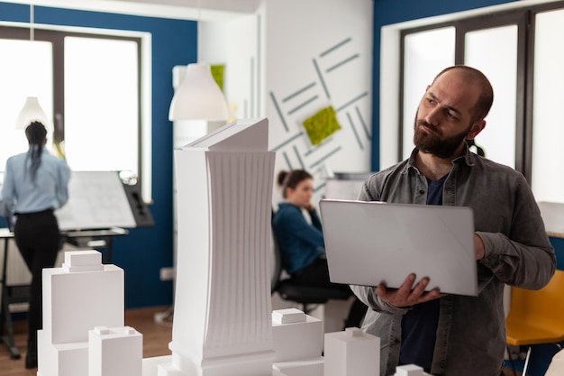 Free Photo architectural inspector holding laptop tilting head looking at maquette of skyscraper in residential project. architect thinking about design improvements to urban planning scale model.