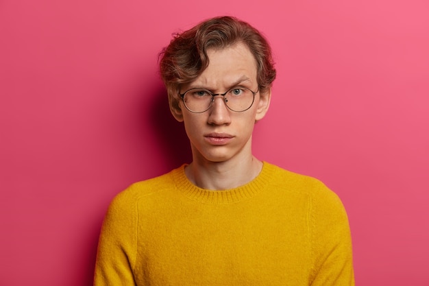 Free photo are you serious? strict indignant man raises eyebrows, has some doubts, being not sure, focused  with doubtful look, wears round transparent glasses and yellow jumper. human face expressions