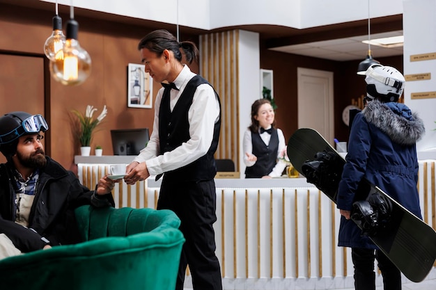 Arrival of female tourist carrying snowboard in hotel lobby for checkin with staff Young waiter serving coffee to man on sofa Winter holiday atmosphere with ski gear Ski resort ensures happy stay