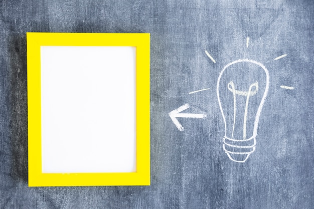 Free photo arrow between white frame with yellow border and light bulb on chalkboard