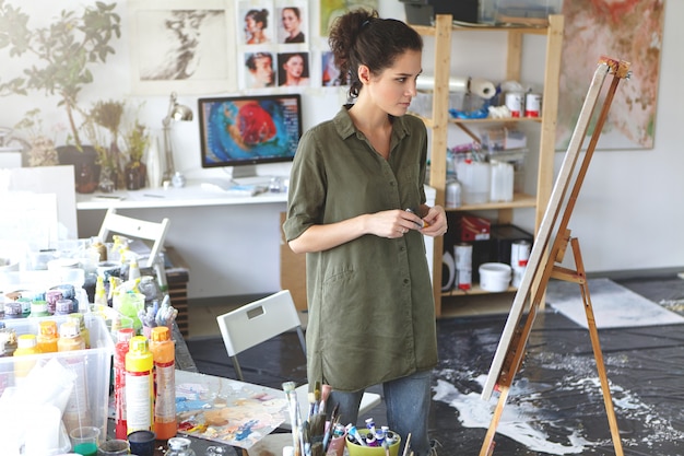 Free photo art and inspiration. indoor shot of hesitant young female artist wearing jeans and khaki shirt standing in spacious workshop interior in front of easel, evaluating painting that she just finished