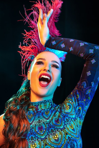 Artist. Beautiful young woman in carnival, stylish masquerade costume with feathers on black background in neon light. Copyspace for ad. Holidays celebration, dancing, fashion. Festive time, party.