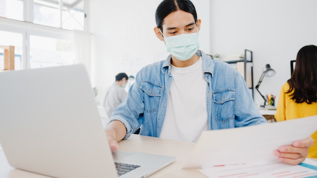 Asia businessman entrepreneur wearing medical face mask for social distancing in new normal situation for virus prevention while using laptop back at work in office. Lifestyle after corona virus.