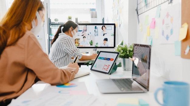 Free photo asia businesspeople wear face mask using desktop talk to colleagues discussing brainstorm about plan in video call meeting in new normal office.