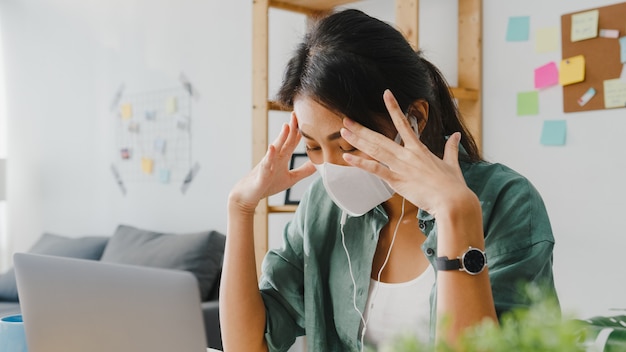 Asia businesswoman wearing medical face mask using laptop talk to colleagues about plan in video call while working from home at living room.